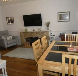 A dining area with a wooden table and chairs next to a sitting area with an armchair a fireplace with a television mounted above and wall art at Aln Cottage in Alnwick