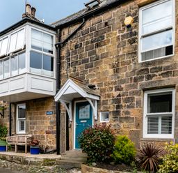 A stone house exterior with a blue front door and white framed windows with plants in front at Begonia House Alnmouth