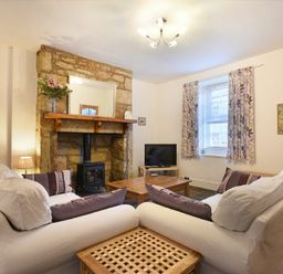 A living room with two white sofas a stone fireplace a television and a window with curtains at Crier Cottage in Alnwick