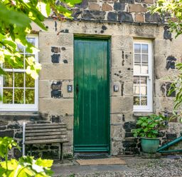 A stone house facade with a green door two white windows a wooden bench and plants at Elliot Cottage in Bamburgh
