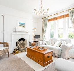 A living room with a white fireplace rattan chair wooden chest and light curtains at Elliot Cottage in Bamburgh