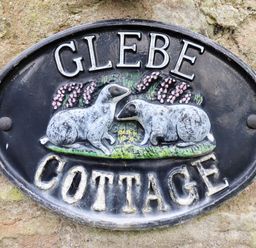 A black oval sign with two white sheep and flowers reading Glebe Cottage attached to a stone wall