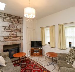 A living room with two sofas a red patterned rug a stone fireplace a small television and cream curtains at Glebe Cottage in Bamburgh