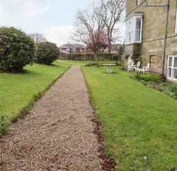 A gravel pathway with bushes and trees beside a stone building with white metal chairs and tables on grass at Glebe Cottage in Bamburgh