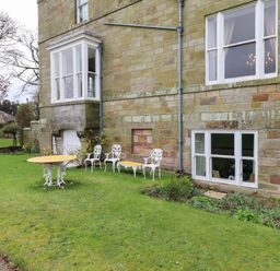 An outdoor area with a stone building wall three white metal chairs one white metal table and a green lawn at Glebe House in Bamburgh