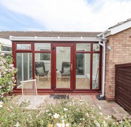 A glass conservatory with chairs inside and a small wooden table chair outside with flowers in the garden at Bedwine Cottage in Beadnell