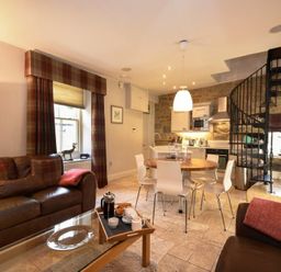 An open living area with leather sofas a glass coffee table a round dining table with chairs a kitchen and a spiral staircase at Coquet Cottage Warkworth