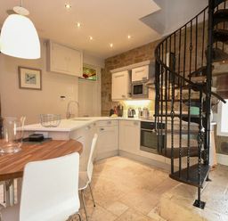 A kitchen with white cabinets a wooden dining table and a black spiral staircase at Coquet Cottage Warkworth