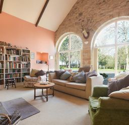 A living room with a bookcase television sofas armchairs wooden coffee table and large arched windows at Craster Tower Coach House Craster