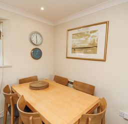 A dining area with a wooden table and six chairs a clock and barometer on the wall a framed picture and a small window with a blind at Dunes Cottage Beadnell