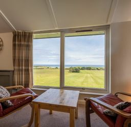 A sitting area with two armchairs and a wooden table by a large window overlooking grassy land and the sea at Fidra in Embleton