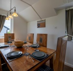 A dining room with a wooden table set with plates and glasses and two windows with curtains at Fidra in Embleton