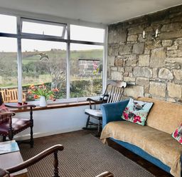 A sitting room with a blue sofa and armchairs next to a large window and stone wall at Leazes Cottage in Rothbury