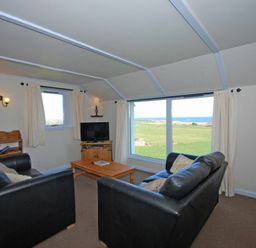 A living room with two black sofas a wooden coffee table a TV on a wooden stand and large windows overlooking a field at Longstone in Embleton