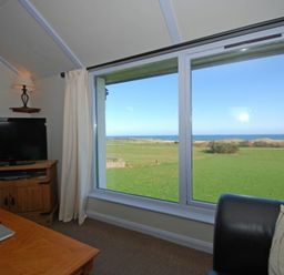 A living room with a television on a wooden cabinet a coffee table a black sofa and large windows showing a green field and sea at Longstone in Embleton