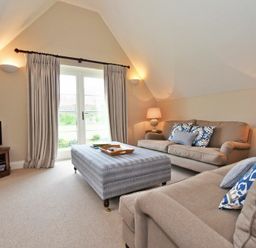 A living room with beige sofas, a large ottoman, lamps, a TV on a wooden stand, and doors with curtains at Sandpiper Bamburgh