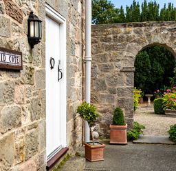 A stone building entrance with a white door and a sign reading Mid Cote with an arched stone gateway leading to a garden with potted plants at Mid Cote Shoreston near Bamburgh