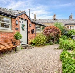 A brick exterior of a cottage with a wooden bench and a gravel path surrounded by green plants and bushes at Nurse's Cottage in Alnwick