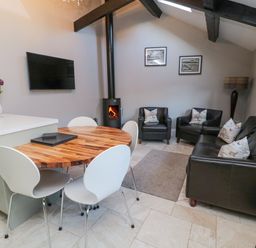 A room with a round wooden dining table and four white chairs a black sofa and two black armchairs with cushions a wood burning stove and a wall mounted TV at Nurse's Cottage in Alnwick