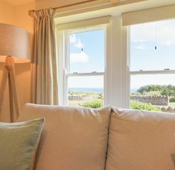 A beige sofa with green cushions near a window with a view of a garden and sea and a floor lamp at Ottoline Cottage in Embleton