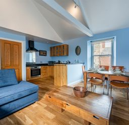 An open plan living room with a blue sofa wooden dining table and chairs and a kitchen with wooden cabinets at Pilots Rest in Alnmouth
