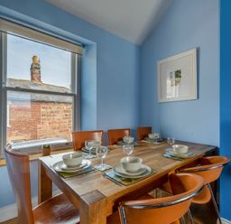 A dining room with a wooden table set for six and brown chairs by a window at Pilots Rest in Alnmouth
