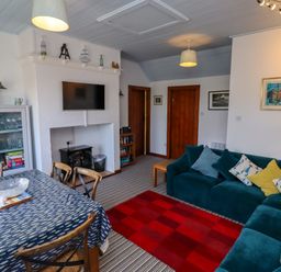 A living room with a blue sofa, wooden chairs around a dining table, a red checkered rug, and a TV mounted above a fireplace at Sand Dune Cottage in Bamburgh