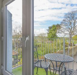 A balcony with a round metal table and chairs overlooking trees and a road at Schooner Court No1 in Alnmouth