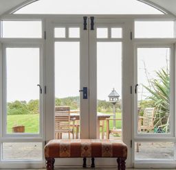 A room with a cushioned bench in front of glass doors leading to a garden with wooden chairs and a bird feeder at Sea Breeze in Newton-by-the-Sea