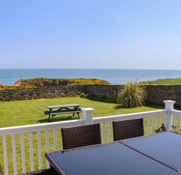 An outdoor patio with wicker chairs and a glass table overlooking a grassy yard with a picnic bench and stone wall by the sea at Seaview Howick in Craster