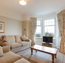 A living room with beige sofas a wooden coffee table a television and windows with floral curtains at Ty y Mor in Seahouses
