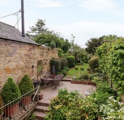A stone wall with patio furniture and potted plants next to a green garden with trimmed bushes at The Book House in Warkworth