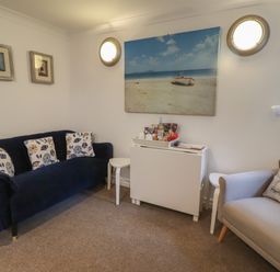 A sitting room with a blue sofa with patterned cushions a beige armchair with a sailboat cushion two small side tables a white cabinet with snacks on top and wall art of a beach and lighthouses at The Little Boathouse in Seahouses