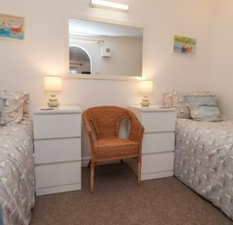 A bedroom with two single beds a wicker chair between two white bedside tables with lamps and a large mirror on the wall at The Little Boathouse in Seahouses