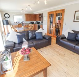 A living area with two black leather sofas and cushions a wooden coffee table with a bowl of decorative items and a lantern a kitchen and dining area with wooden cabinets a table and chairs and a wall clock at Tree Corner in Beadnell