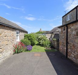 An outdoor space with stone buildings on either side a paved area and a grassy path with bushes and flowers at Wynding Down in Bamburgh