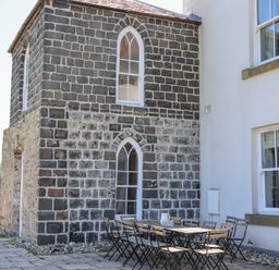 An outdoor patio area with a long wooden table and folding chairs next to a stone building with arched windows at The Lookout Embleton