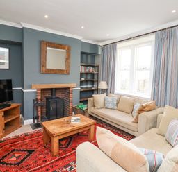 A living room with beige sofas a wooden coffee table a patterned red rug a television on a wooden stand a fireplace and striped curtains at The Hollies in Bamburgh