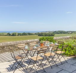 An outdoor patio with wooden tables and chairs overlooking fields and the sea at No6 Sea Lane in Embleton