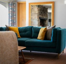 A living room with a teal sofa and yellow and brown pillows with a large mirror reflecting a stone wall at The Arches in Bamburgh