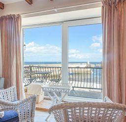 A seating area with wicker chairs and a small white table facing a balcony with a view of the harbor and sea at Farne Lookout in Seahouses