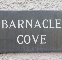 A grey rectangular plaque with the text Barnacle Cove mounted on a white textured wall