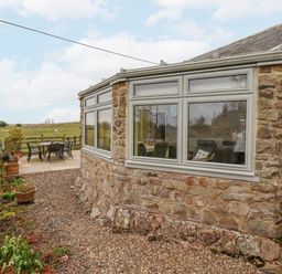 A stone exterior of a house with large windows and a patio table and chairs in a field at Adderstone Cottage in Bamburgh
