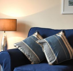 A blue sofa with two striped pillows and a table lamp on a side table at Beachcomber Apartment in Bamburgh