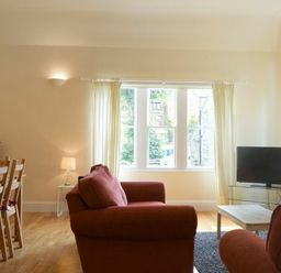 A living room with red sofas a wooden dining table and chairs a TV on a glass stand and large windows with curtains at Decked Out in Alnmouth