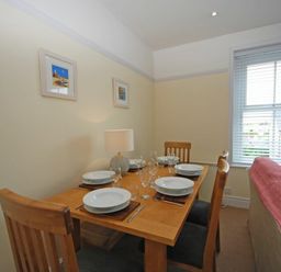 A dining area with a wooden table set for six with plates glasses and cutlery next to a window with blinds and a sofa with a red throw at Coastal Haven in Seahouses