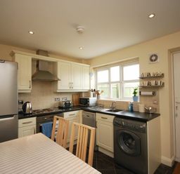 A kitchen with white cabinets black countertops a silver refrigerator a washing machine a dining table with wooden chairs and a window at House Martins Beadnell