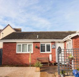 A small single-story brick house with a white door windows a wooden bench a hanging plant a garden trellis and a blue ceramic pot at Arcadie in Amble