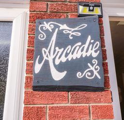 A black wooden sign with white lettering that reads Arcadie mounted on a red brick wall near a window and a door at Arcadie in Amble