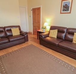 A living room with two brown leather sofas with yellow pillows a wooden door a side table with a lamp and flowers and a rug on a wooden floor at Riverside Cottage in Warkworth